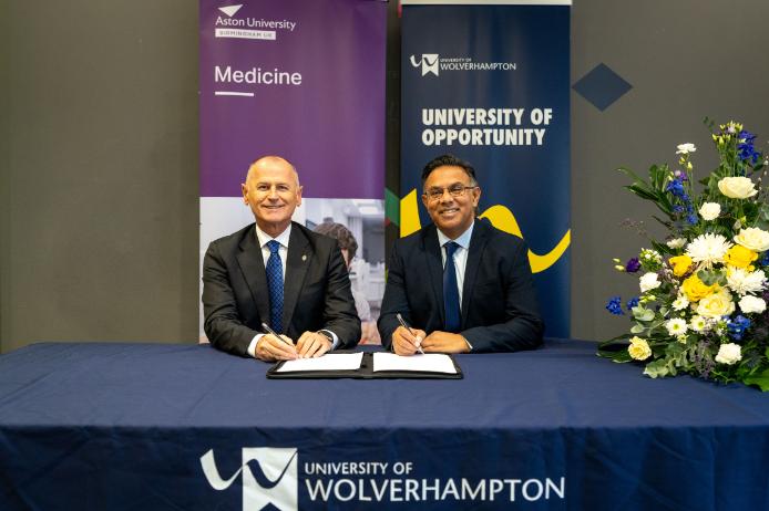 Professor Aleks Subic, Vice-Chancellor and CEO of Aston University and Ebrahim Adia, Vice-Chancellor of University of Wolverhampton sit at a table with the two University's banners behind them. They hold a pen ready to sign the agreement