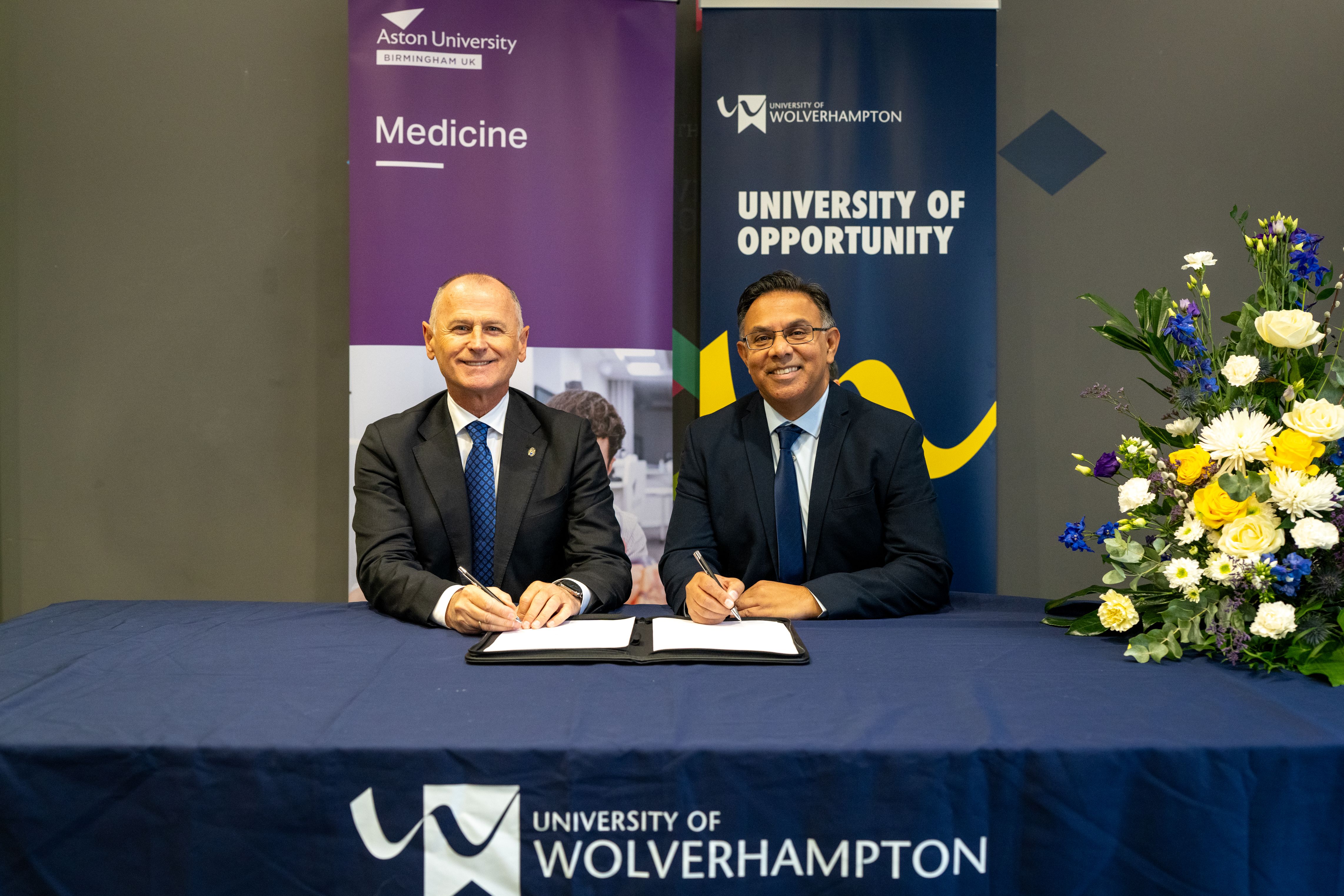 Professor Aleks Subic, Vice-Chancellor and CEO of Aston University and Ebrahim Adia, Vice-Chancellor of University of Wolverhampton sit at a table with the two University's banners behind them. They hold a pen ready to sign the agreement
