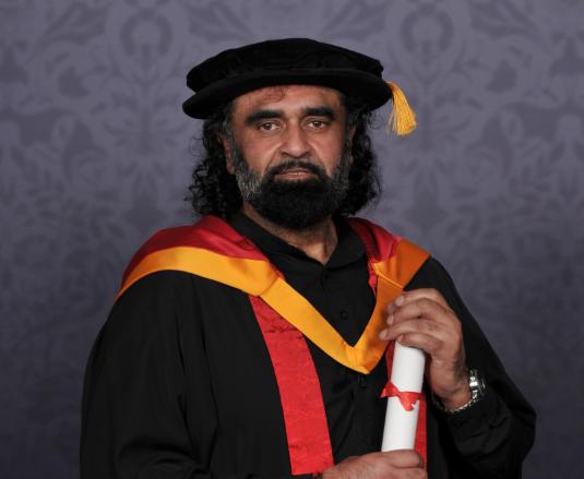 Local sports hero Ranjit Singh poses for a professional graduation photograph dressed in University of Wolverhampton yellow and orange robes and cap. He holds a scroll between his hands.