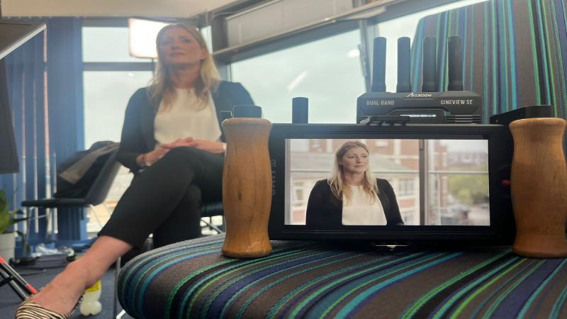 Rebecca Flanigan sits in a chair with a camera in front of her. She is blurred out in the background but a crisp image of her on the camera's display is on a chair in the foreground.
