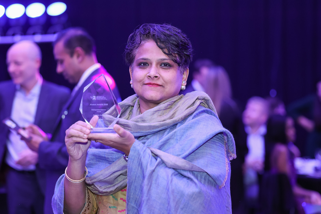 Rezwana is dressed in yellow dress with blue shawl. She holds up her award for the camera. Behind her are people celebrating at the 2025 Alumni Awards celebration.