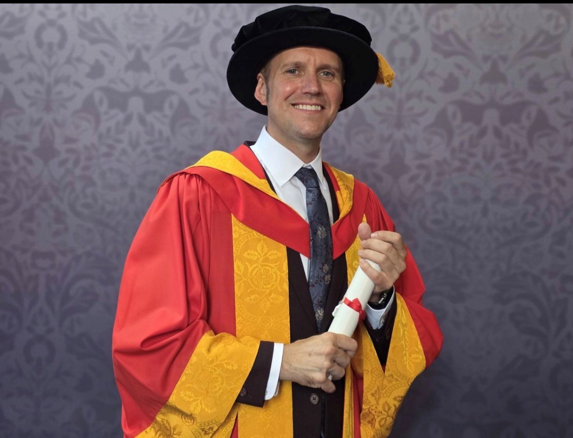 Rob poses for a professional graduation photo dressed in graduation robes in University of Wolverhampton colours, yellow and orange. He holds a scroll between his two hands and smiles for the camera.