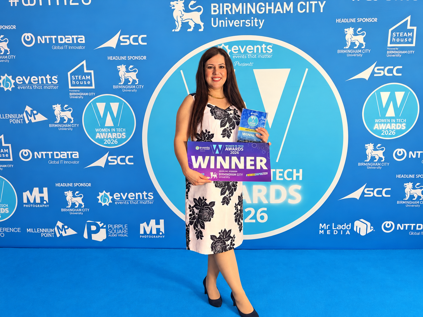 Professor Roya Rahimi holds her award. She's wearing a white dress with black flowers and stands in front of a backdrop covered in sponsor logos.