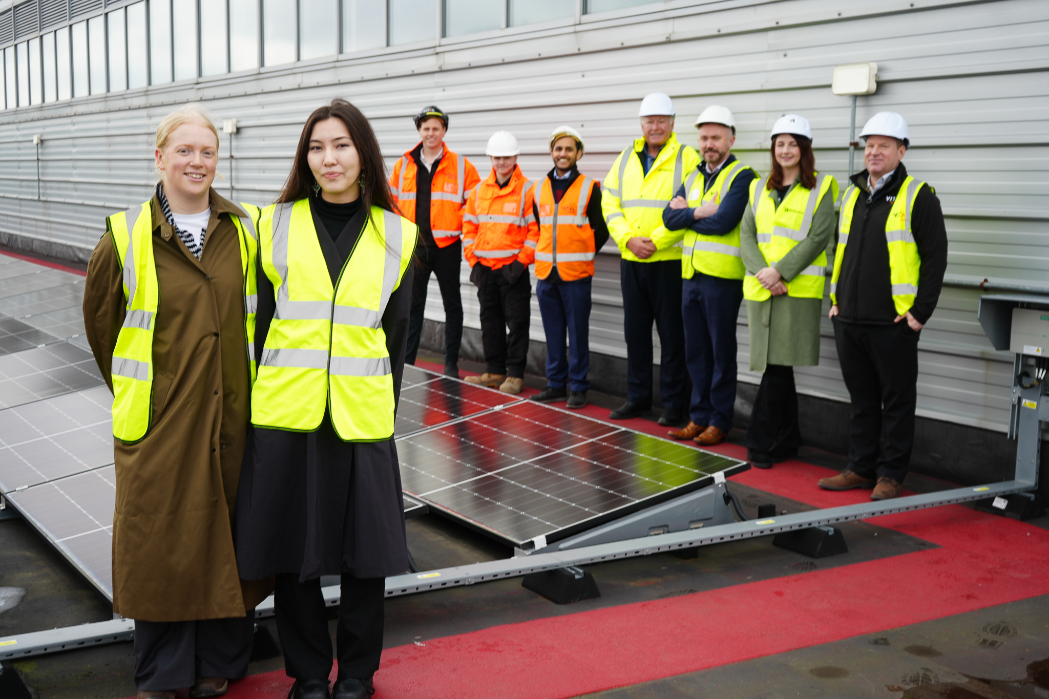Two representatives from Salix Finance are picture in the foreground wearing high viz jackets in front of solar panels on a roof at Walsall Campus. Pictured behind them are representatives from the University of Wolverhampton and the project contractors VitalEnergi