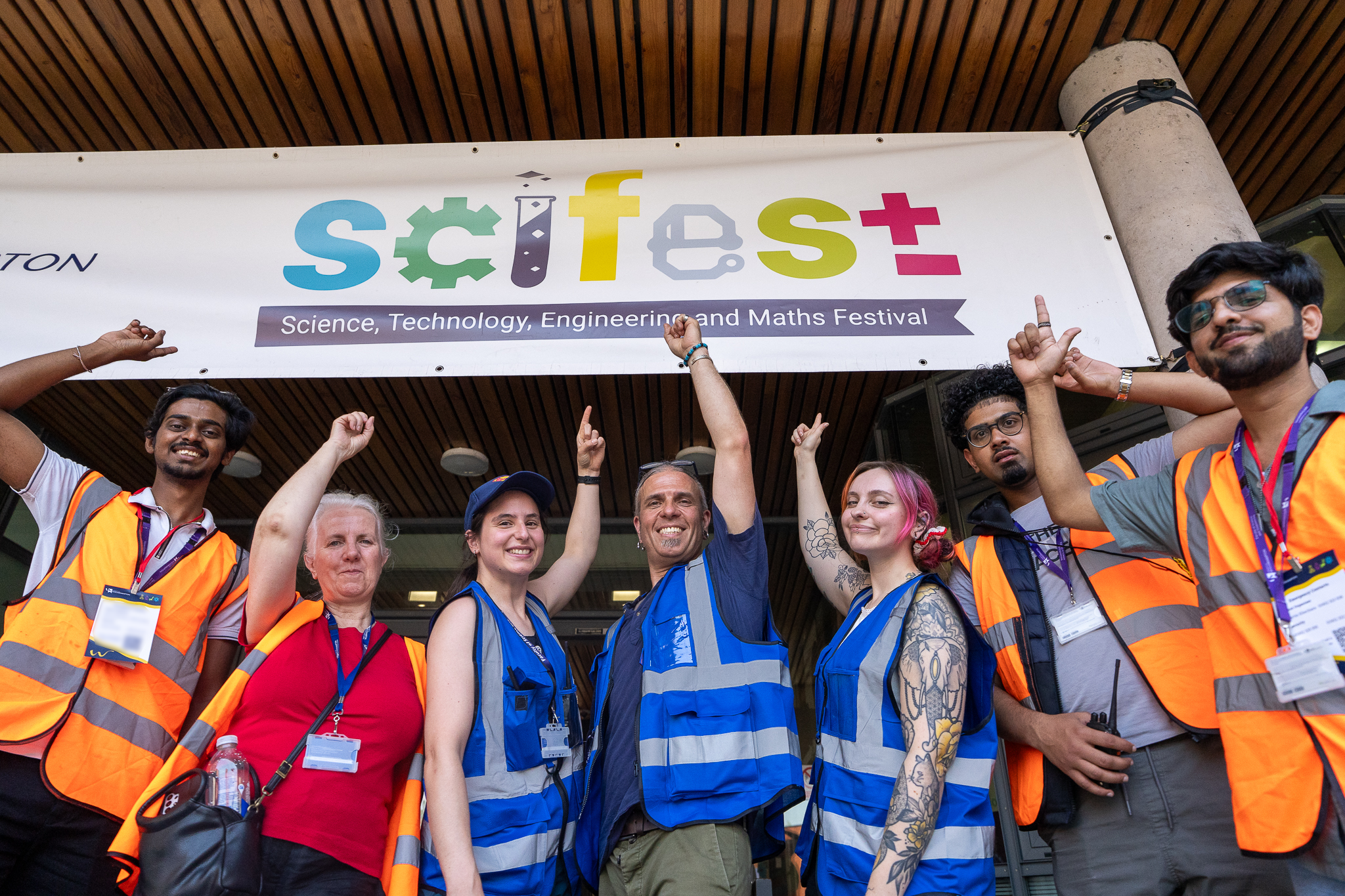 A picture of four men and three women in a line. They are wearing hi-viz jackets in blue and orange. The are all pointing up at a sign above them saying SciFest.