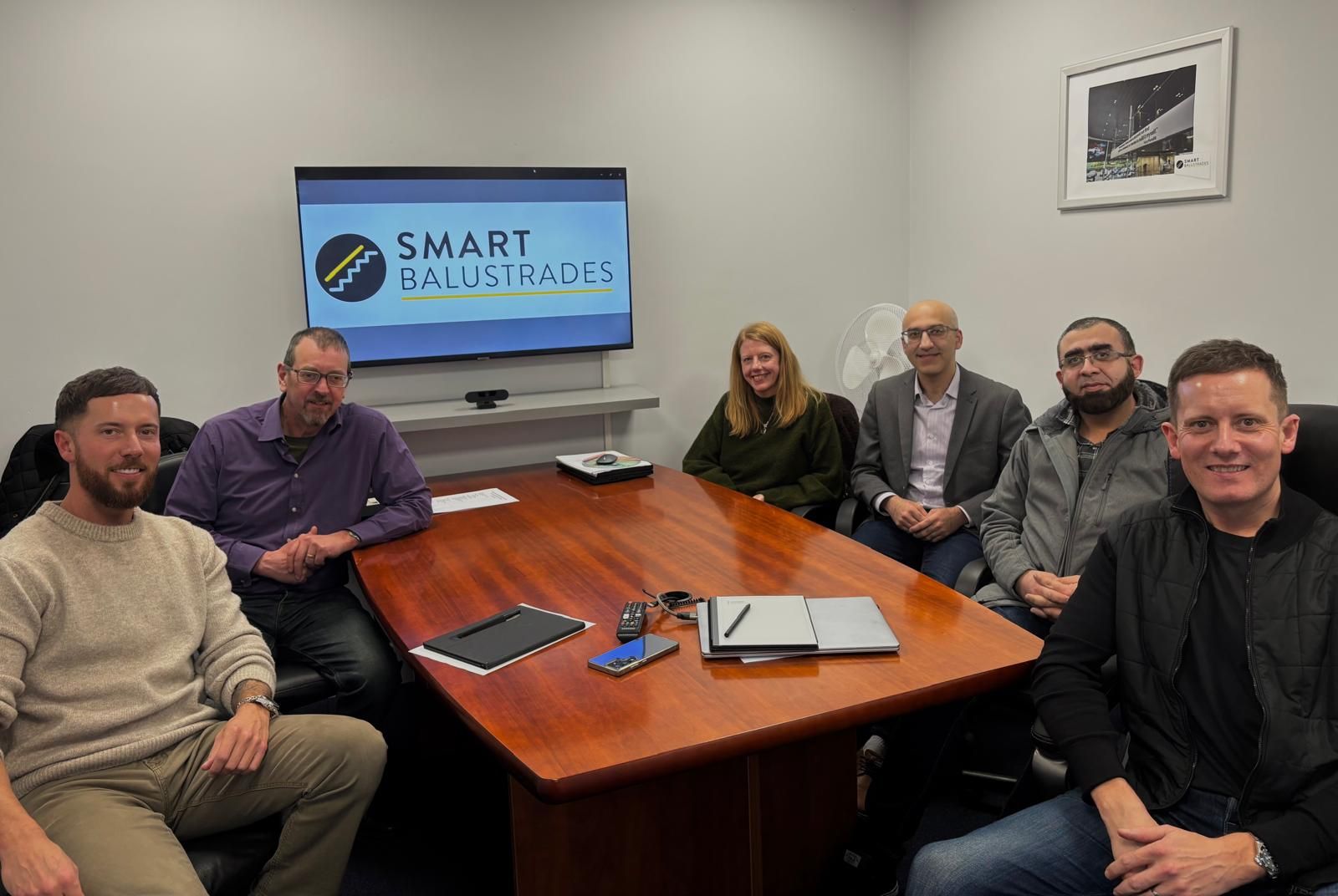 Six individuals from the University of Wolverhampton and SMART Balustrades sit around a table. There is a screen behind displaying the SMART Balustrades logo.