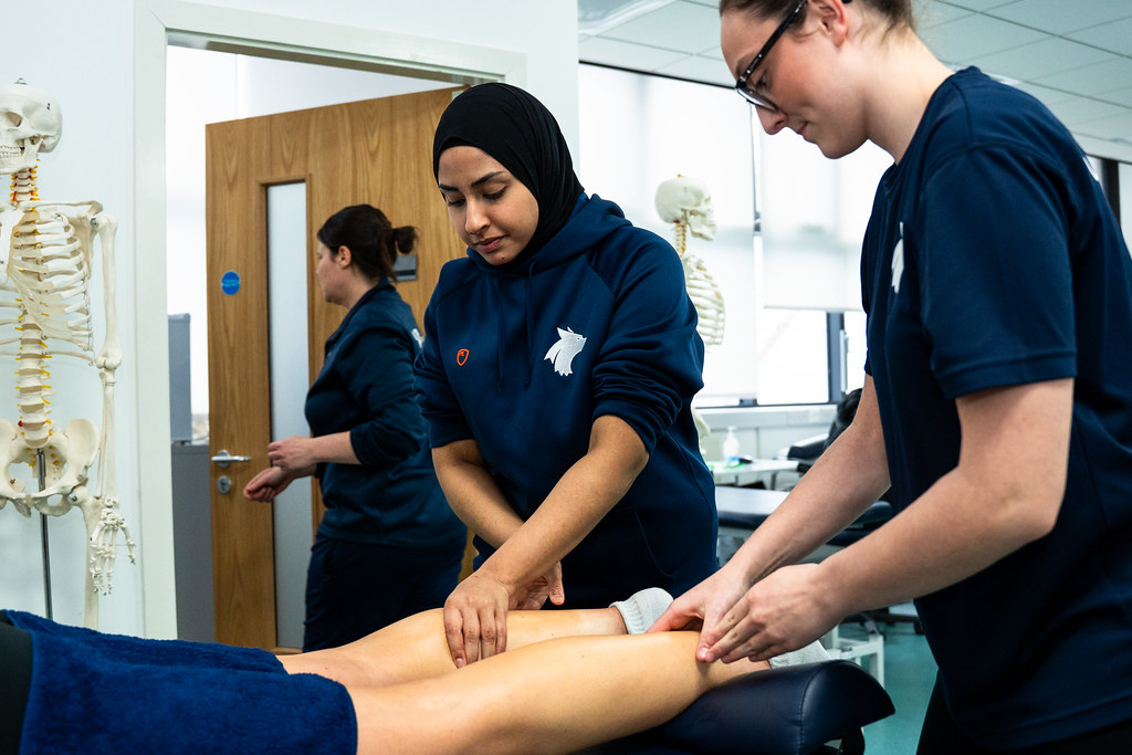 Two students wear Wolverhampton polo shirts in navy blue. They lean over a therapy table and massage someone's legs