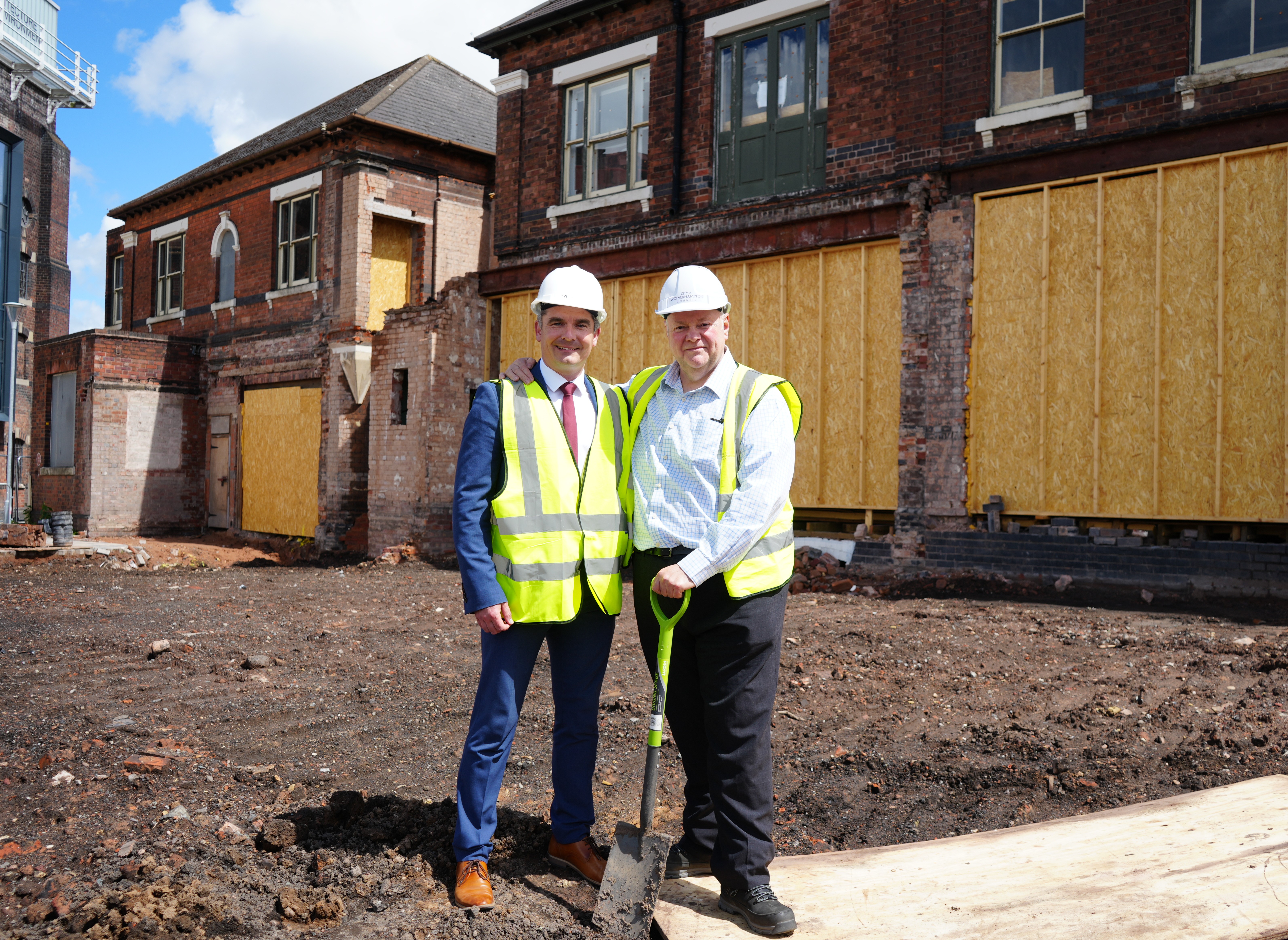 Dr Peter Cross (left) wears a suit, hi-viz waistcoat and white hard hat and Cllr Stephen Simkins (right) wear trousers and shirt with hiz-viz waistcoat and white hard hat. Stephen holds a spade in his right hand. They are standing on mud at the site of the demolished building at Springfield Campus.