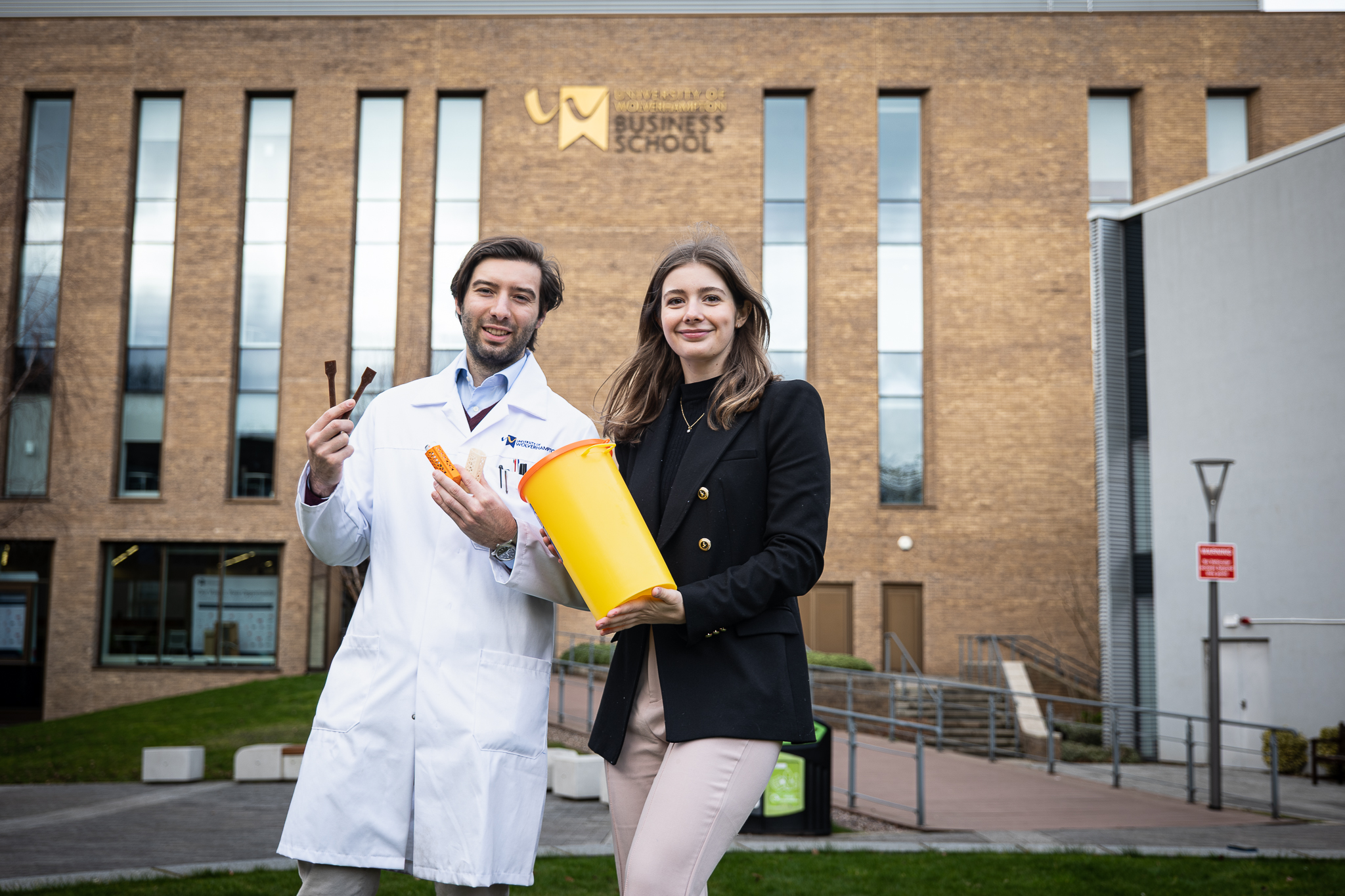 The company founders Olivia Simpson and Dr. Mattia Parati show a product made with seaweed based material. Dr Parati wears a white coat and holds the product in his hands and Olivia Simpson holds a yellow receptacle. They are outside the University's business school.
