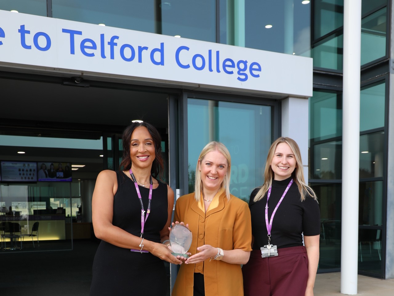 Three women stand in line outside Telford College. They are dressed in business attire and the women in the centre hands an award to the woman on her right hand side.