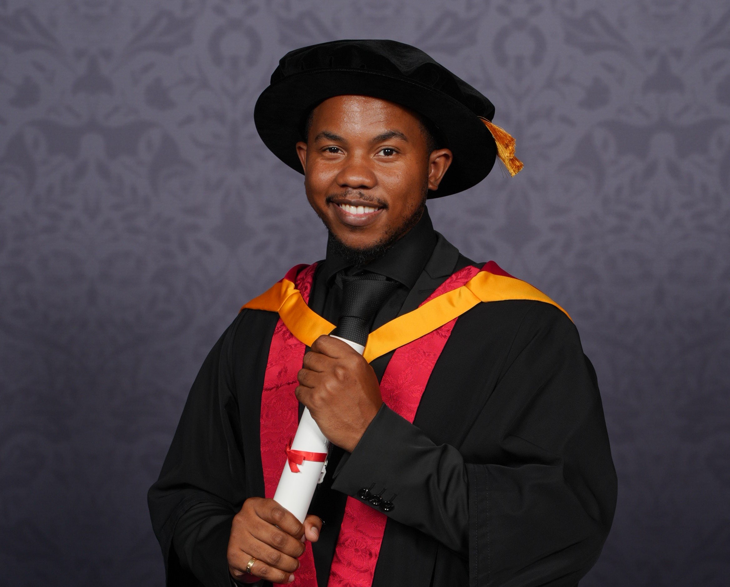 Theo poses for a professional graduation photo dressed in graduation robes in University of Wolverhampton colours, yellow and orange. He holds a scroll between his two hands and smiles for the camera.