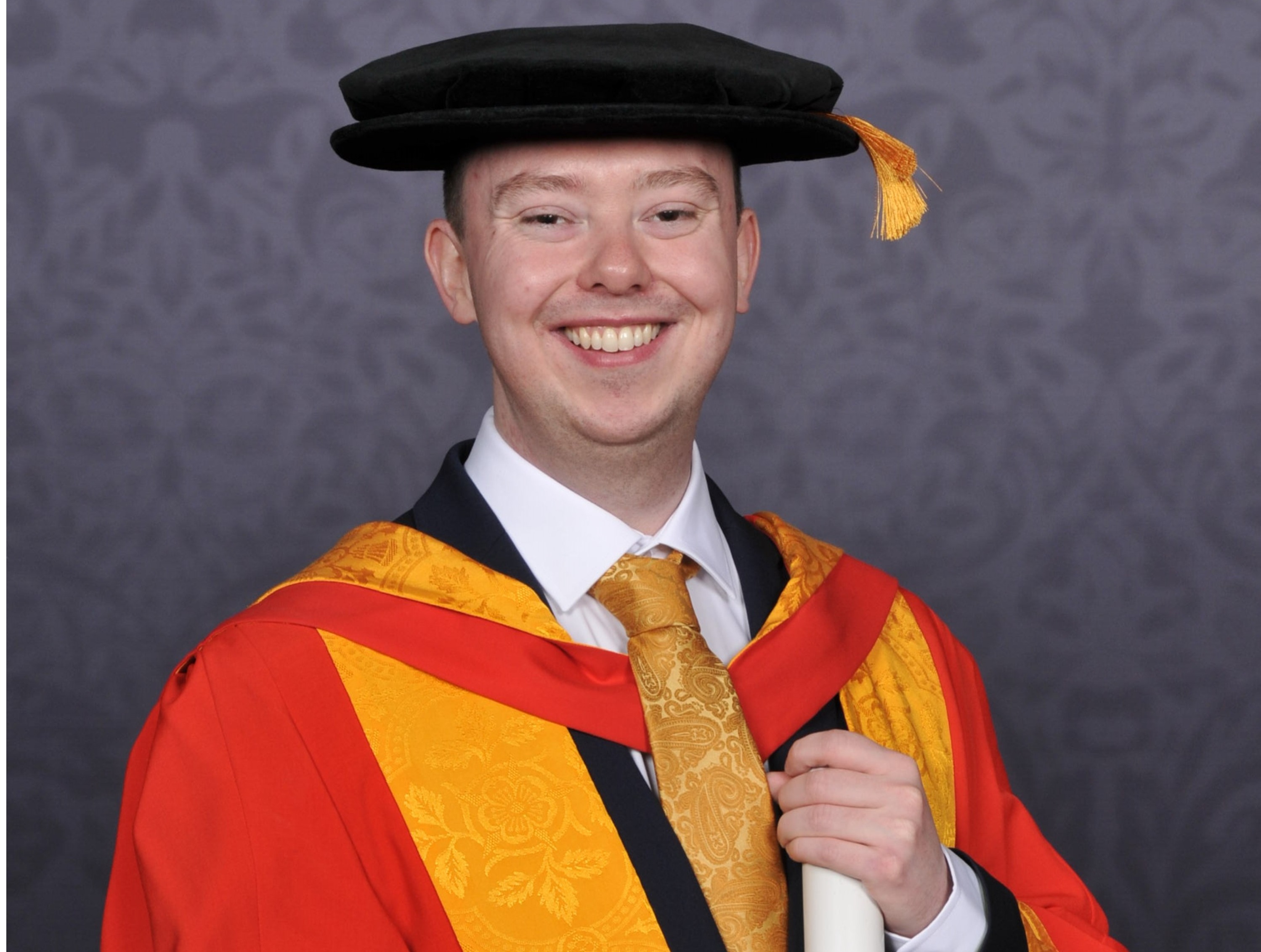 Walker Darke poses for a professional graduation picture, holding a scroll and wearing University of Wolverhampton's signature yellow and orange gown