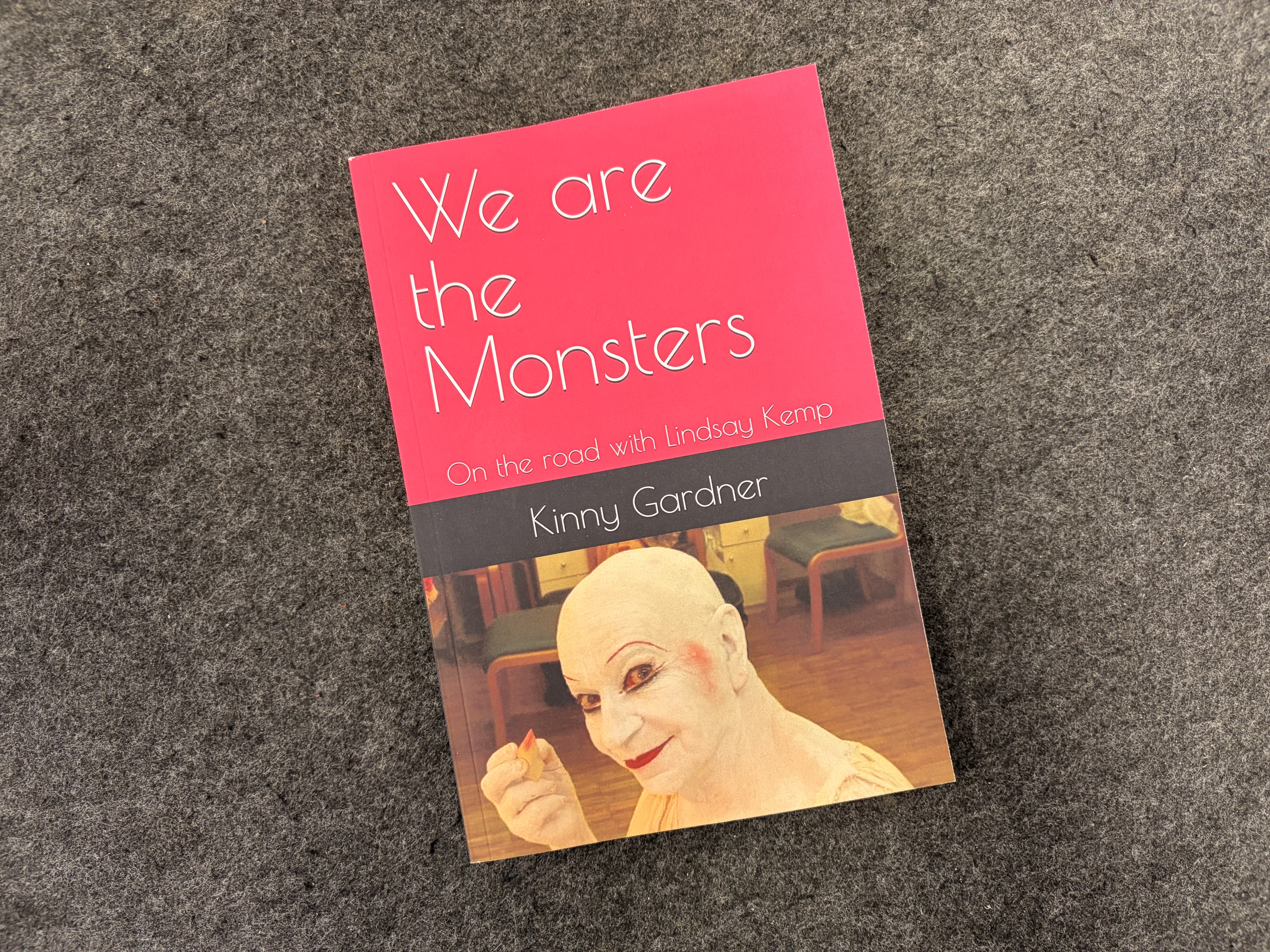 A photo of a book cover - We are the Monsters. The book is red with a face painted in white face paint, looking like a clown
