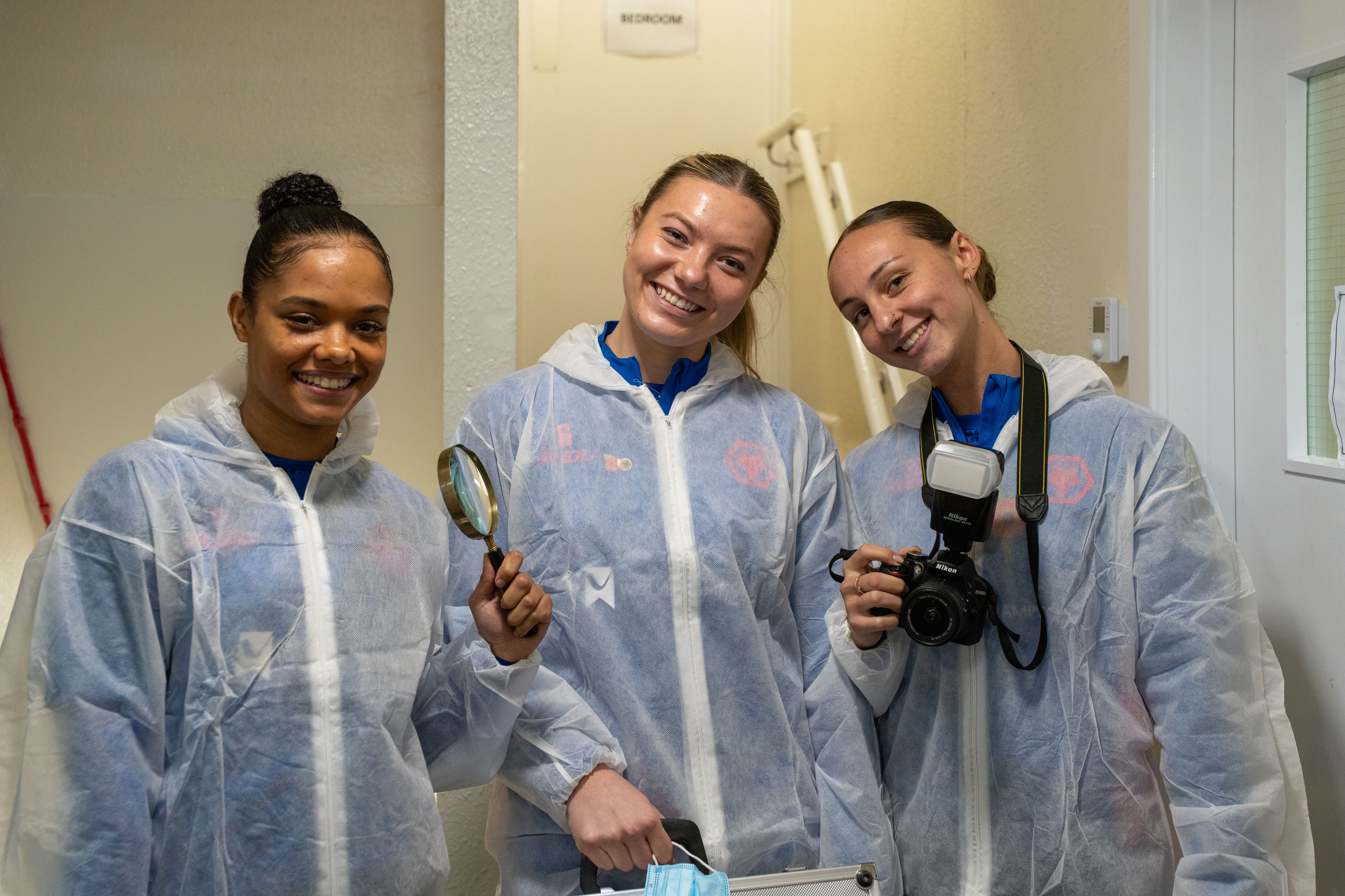 Anna Grey, Lily Simkin and Louanne Worsey dressed in overalls, ready for their investigation at Locard House. They hold a magnifying glass and a camera.