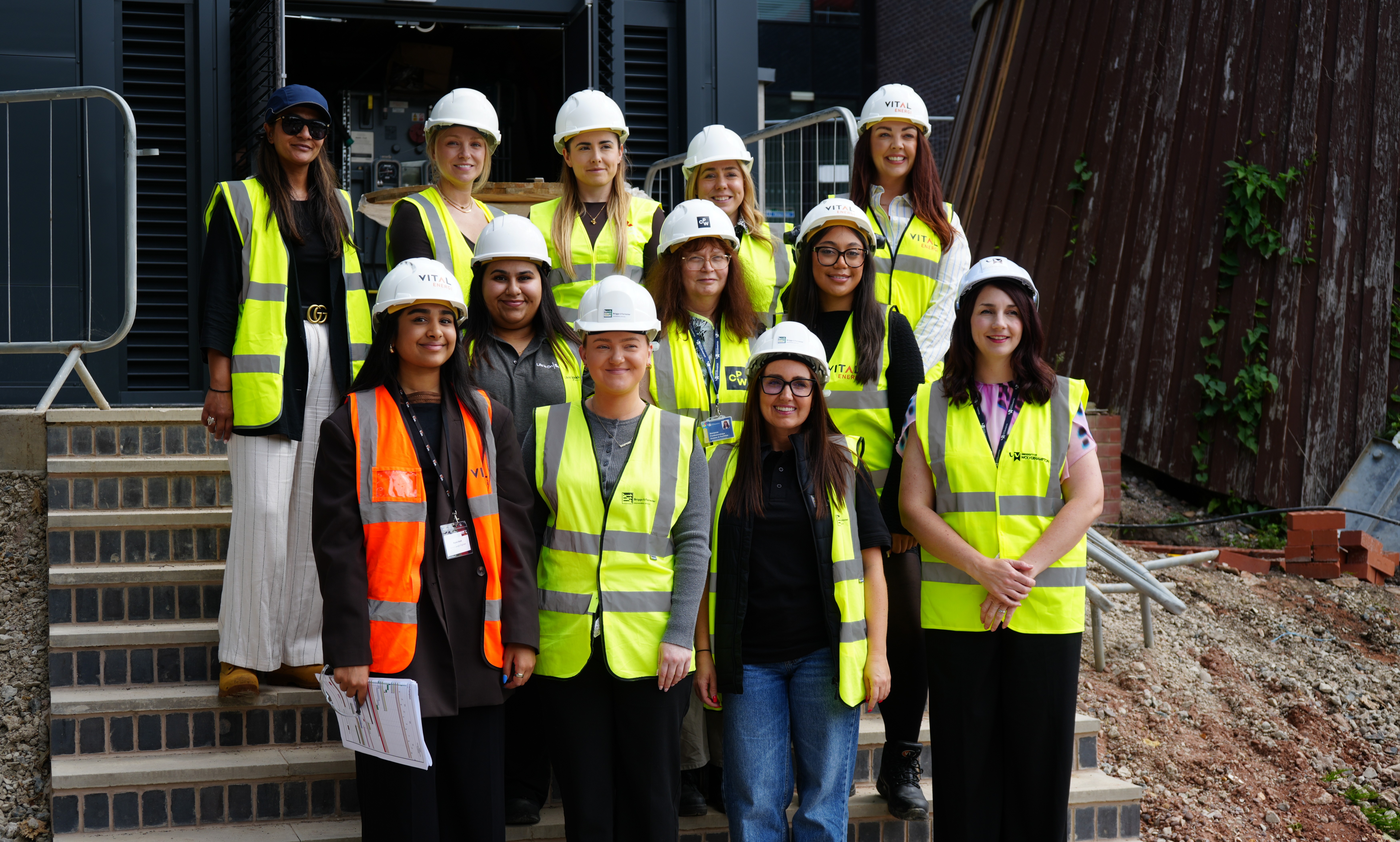 Twelve women stand on the steps of a site at Walsall Campus as part of a site tour. They wear casual clothes, hard hats and hi-viz waistcoats and smile for the camera.