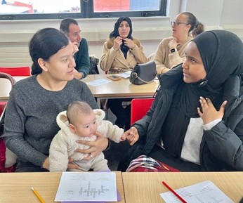 Two women talking while sat at desks with worksheets and pens in front of them. The woman on the left is holding a baby on her lap. Behind them is a man and two women also talking at a desk with worksheets on the desk.