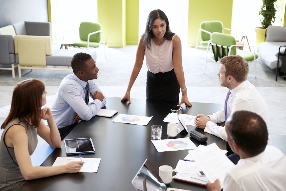 Header image of a boss at a table with her colleagues talking to her