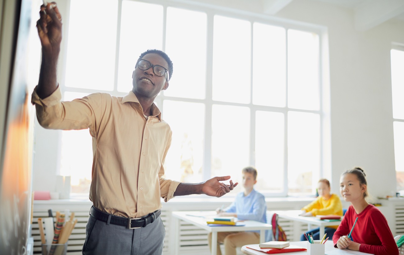 Header image of a teacher writing on a blackboard while students watch at desks