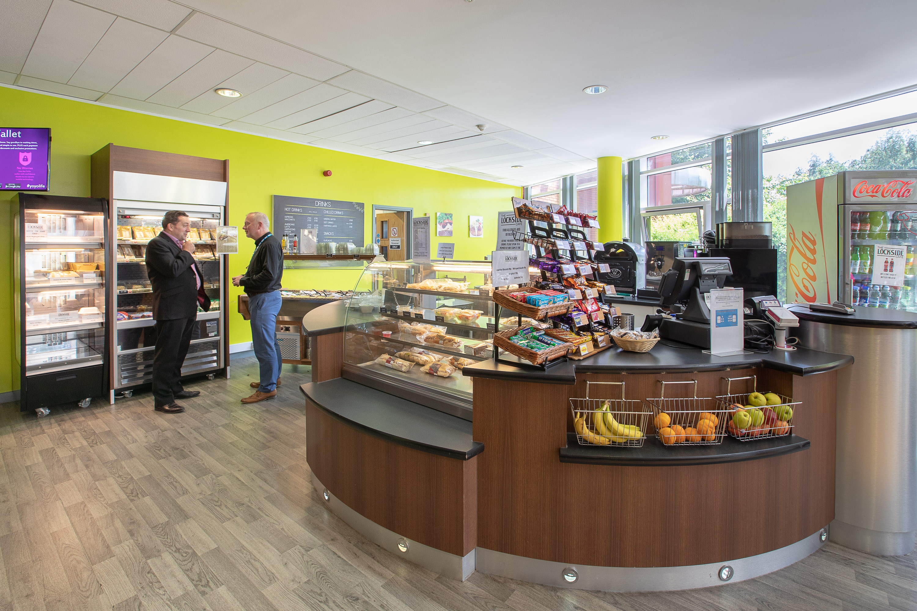 Two people standing inside the Lockside Café next to food refrigerators, opposite a counter with cooked food and fresh fruit on display, a drinks vending machine and a menu blackboard visible in the background
