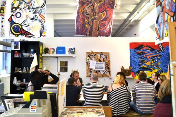 Display of various art pieces by students in the School of Art, with students sitting around a table as pictures hang from walls and the ceiling of the room