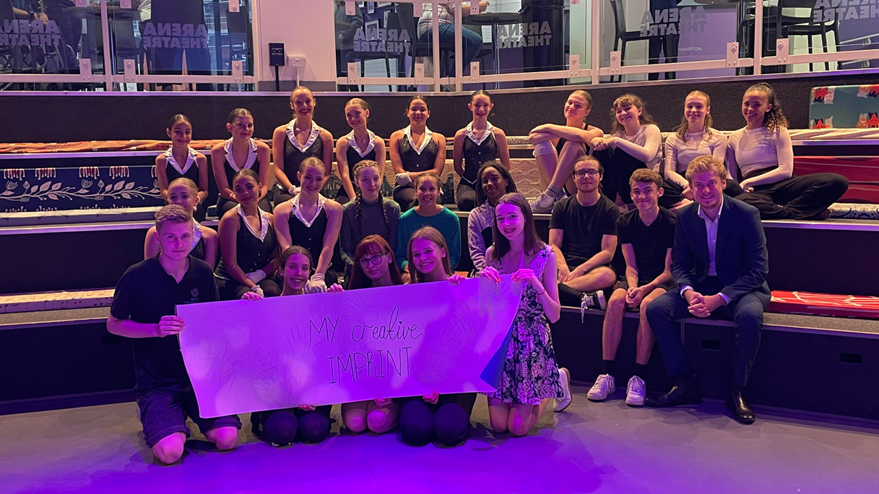 Group photo of young people, aged between 11 and 15, taken in the Tilson Studio within the Arena Theatre. They are sat together on the ampitheatre steps holding a banner that reads: 