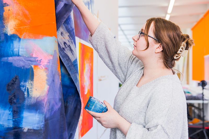 Female student working on a large abstract painting  of mostly orange, blue and purple