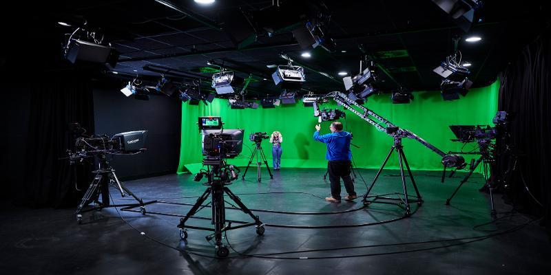 Two people using the large TV Studio in the Wolverhampton Screen School - one male one female. there is a large green screen in the background and several cameras and a full lighting rig in the foreground of the image. The figures are in the background, one of whom is barely visible.