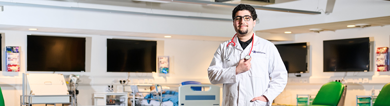 medical student in white coat, standing in mock ward with stethoscope around his neck.