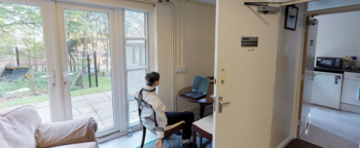 Forensic Science Crime Scene House at City Campus, the interior of a living room wiht a sofa and a dummy of a man sitting at a work desk. There is a door open showing the view into a kitchen.