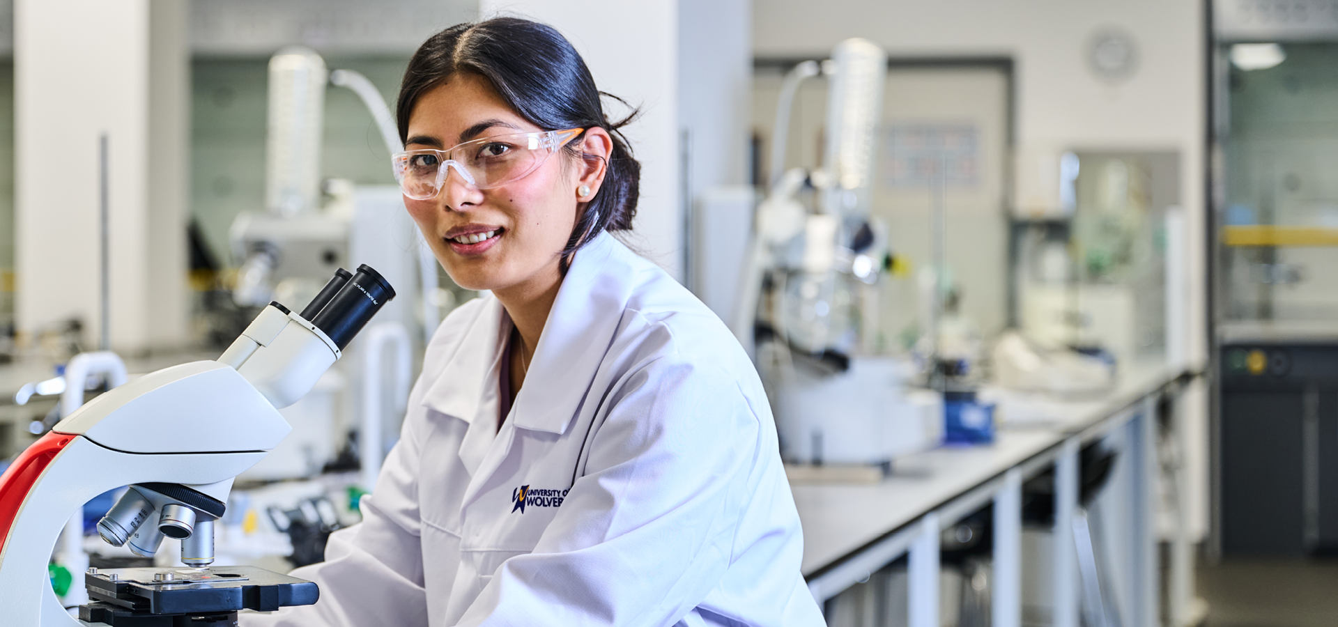 Pharmaceutical Science student in City Campus science lab wearing a white lab coat and safety goggles operating a microscope on the work surface.