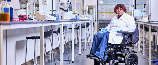 Student in wheel chair wearing white lab coat and blue gloves sitting in pharmacy labs at City Campus