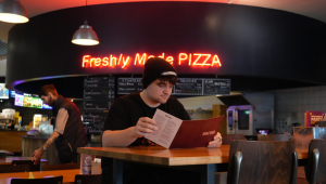 A student examining the menu in the Light cinema pizza restaurant, a neon sign in the background reading FRESHLY MADE PIZZA