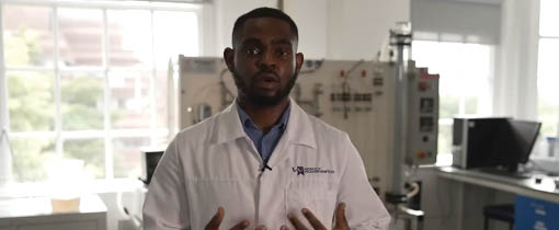 Student standing in a chemical engineering lab and wearing a lab coat and safety goggles.