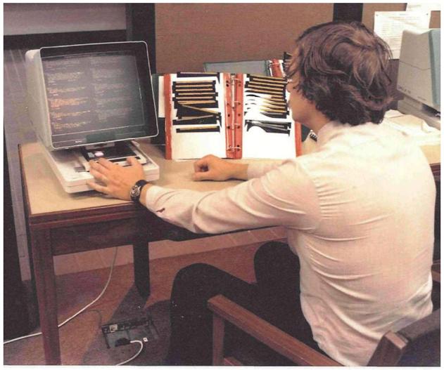 Historical photograph of a person sitting at a desk using a rudimentary computer.