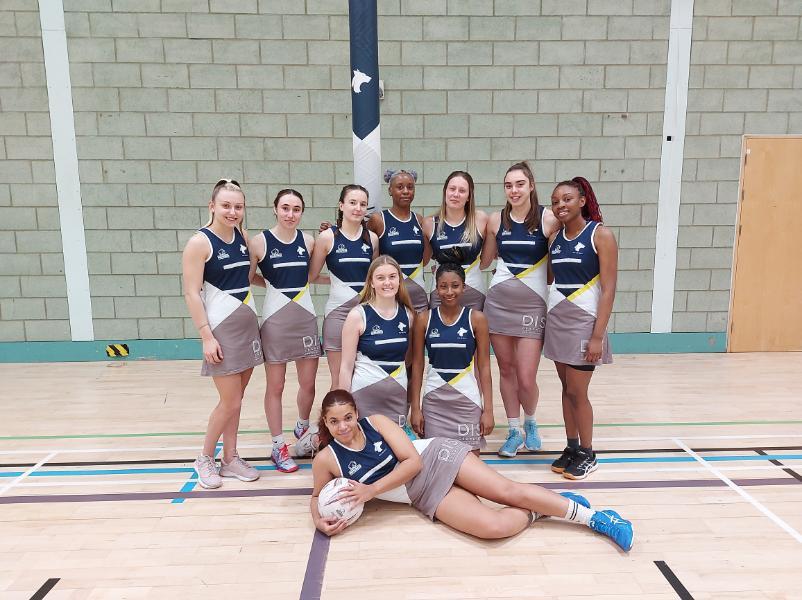 The University of Wolverhampton Womens Netball team, some members standing around others on their knees and one on the floor of the sports hall with a netball